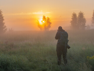 A photographer stands in a misty field at sunrise, capturing the golden morning light filtering through distant trees and fog © Александр Арендарь