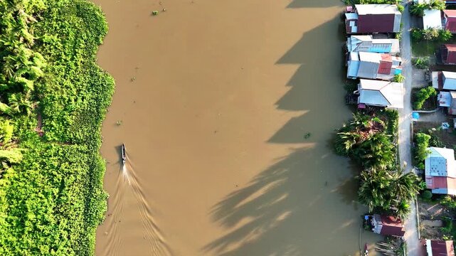 Aerial view of a small boat navigating the river with lush vegetation and riverside houses in Banjar Regency, South Kalimantan, Indonesia.