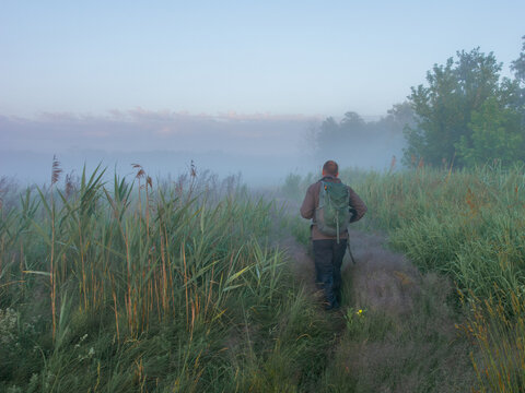 A man with a backpack walks down a grassy path surrounded by reeds into a thick fog during early morning sunrise