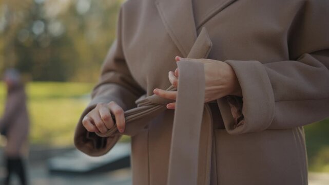 Close up of woman tying belt on her brown coat in outdoor park setting. focus remains solely on hands and fabric manipulation with soft natural light illuminating scene. Background shows
