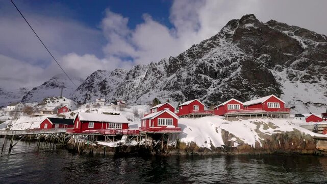 View of traditional red wooden rorbuer cabins built on stilts over the water against a backdrop of snow-covered mountains in A, Nordland, Norway.