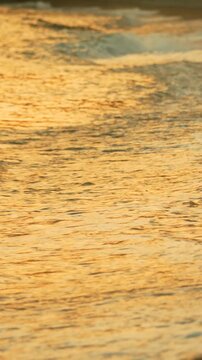 Amber sea surf glides over dark wet pebbles on a sandy beach during twilight, revealing rhythmic natural cycles. Static vertical camera shot.