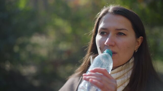Close up of woman drinking water from plastic bottle in outdoor setting. subject holds clear partially filled container with both hands while sunlight illuminates her face and long hair