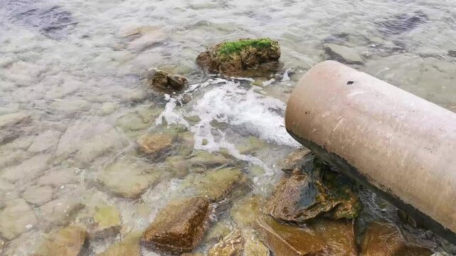 Wastewater pipe discharging water onto rocky shoreline in daylight