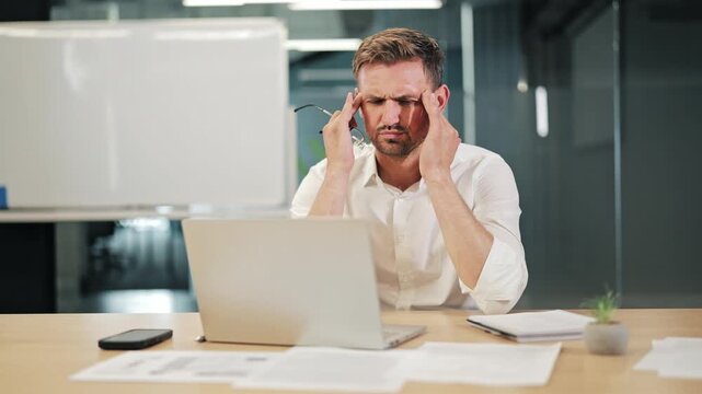 Professional at workstation experiences intense headache. Business demands cause significant stress, eye strain. Overworked individual shows signs exhaustion, pain.