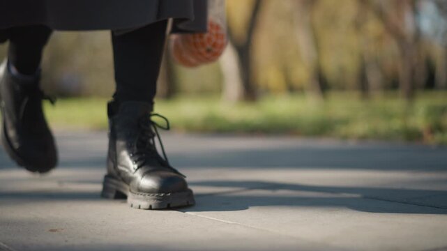 Close up of person feet in black leather boots walking on pavement holding orange mesh bag with fruit. scene shows consistent outdoor environment with greenery and buildings in soft daylight