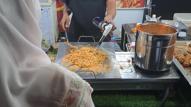 Street food vendor torches creamy macaroni cheese on hot griddle with flames at night festival. Cheesy pasta, blowtorch cooking. PAMEKASAN, INDONESIA - JUNE 08, 2026
