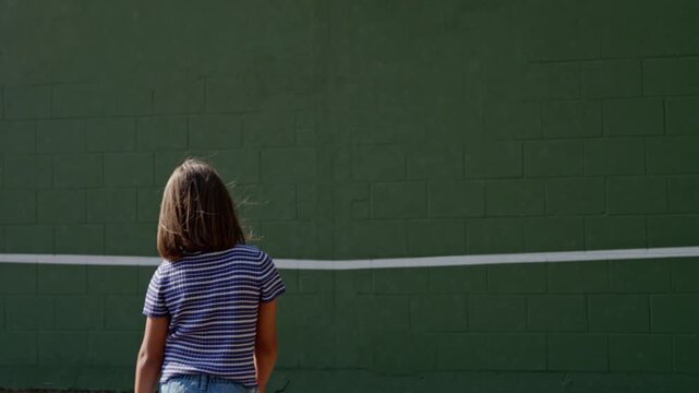 Teen girl practicing tennis against green wall, body turning through strong swing with striped shirt, short hair, rear view, outdoor court texture, focused training energy