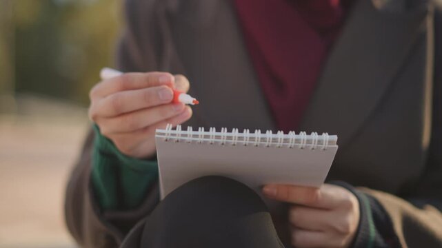 Close up of woman holding and writing in spiral notebook with pen focusing on her hands and open page. scene shows soft outdoor lighting as woman interacts with stationery items capturing