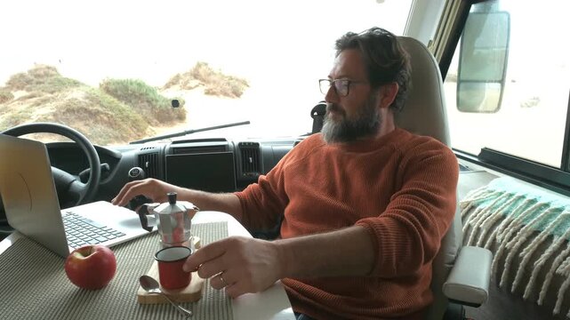 Middle-aged man working on a laptop and drinking coffee inside his motorhome, enjoying the freedom of a digital nomad lifestyle and an alternative way of living on the road