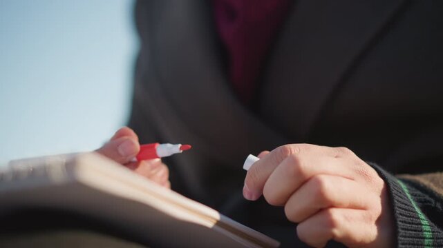 Close up of hands holding red pen over open notebook fingers poised to write. Notebook pages visible with soft lighting and clear texture detail. Background blurred emphasizing focused