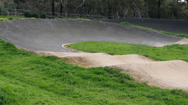 A steep asphalt banked turn leading into dirt rhythm jumps on an outdoor BMX track. Sealed cornering surfaces and packed dirt straightaways with rhythm rollers and mounds. Melbourne, Australia