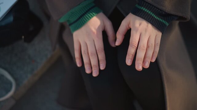 Close up of hands resting on lap with fingers relaxed and slightly apart. Hands are positioned centrally in frame showing natural skin texture under soft lighting. Background includes