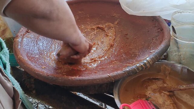 Close up of hands grinding Madura petis, peanuts, chili, tomato in clay mortar for authentic rujak cingur. Traditional Indonesian street food preparation, East Java culinary culture, spicy sauce.
