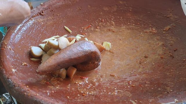 Close up of hands grinding Madura petis, peanuts, chili, tomato in clay mortar for authentic rujak cingur. Traditional Indonesian street food preparation, East Java culinary culture, spicy sauce.