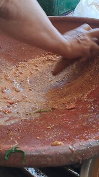 Close up of hands grinding Madura petis, peanuts, chili, tomato in clay mortar for authentic rujak cingur. Traditional Indonesian street food preparation, East Java culinary culture, spicy sauce.