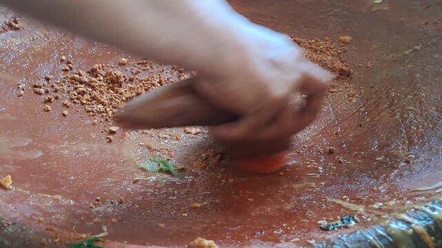 Close up of hands grinding Madura petis, peanuts, chili, tomato in clay mortar for authentic rujak cingur. Traditional Indonesian street food preparation, East Java culinary culture, spicy sauce.