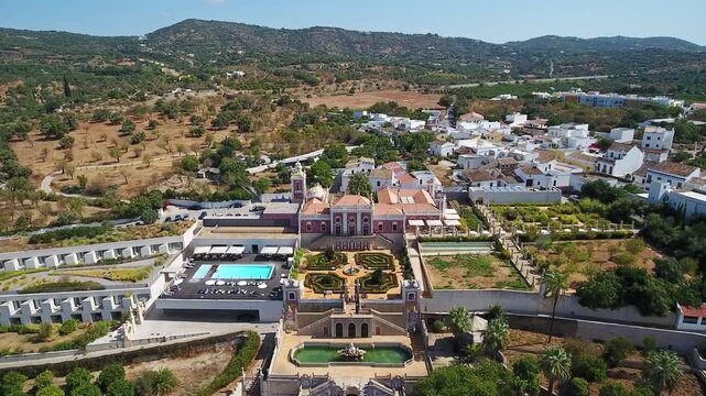 Aerial view flying towards the historic pousada palacio de estoi, revealing its magnificent rococo architecture, french style gardens, and a modern luxury hotel wing with a swimming pool