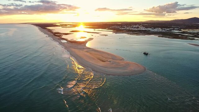 Golden sunrise light reflects on the water as an aerial view captures a dredging boat working near a sand spit in ria formosa natural park, creating a channel in fuseta, algarve, portugal
