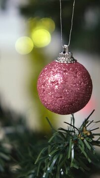 Glittering dusty rose Christmas bauble hanging above pine branch. Vertical format, textured holiday bulb swinging on thread as cozy winter aesthetic scenery. Gen-Z cozy design vlogging