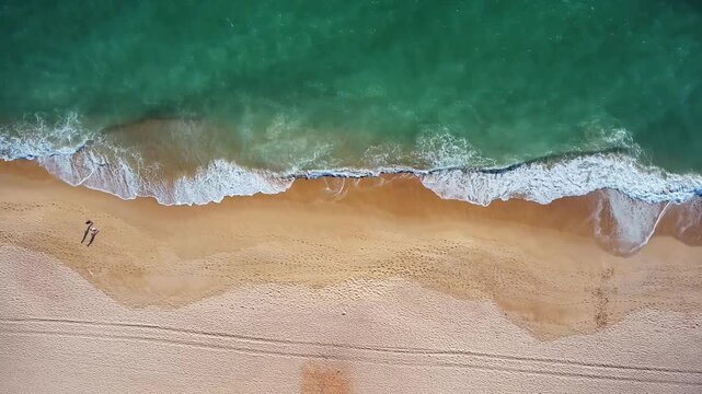 Aerial top down view of a young couple walking along a pristine sandy beach as turquoise ocean waves gently wash ashore, creating a serene and romantic atmosphere for a perfect vacation getaway