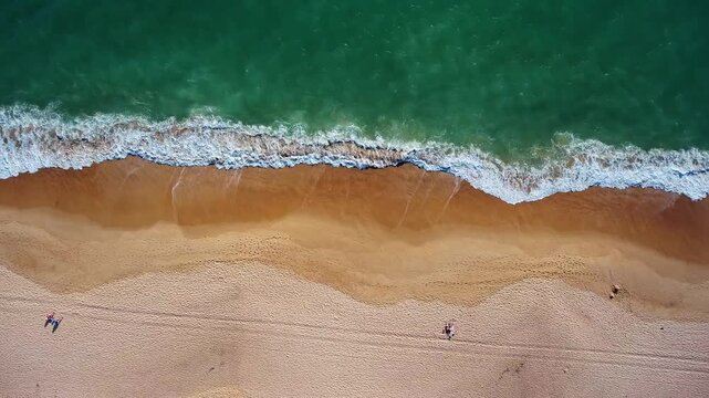 Majestic aerial top down perspective capturing turquoise ocean waves gently rolling onto a vast, golden sandy beach while a few people enjoy a relaxing stroll along the picturesque shoreline