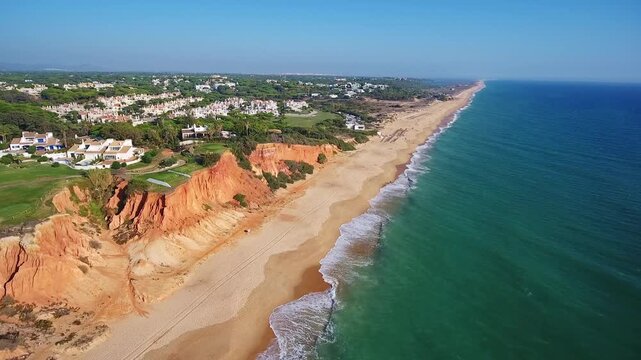 Beautiful aerial footage showing the stunning algarve coastline with its iconic red cliffs, sandy beaches, and luxury resorts in vale de lobo, portugal, under a clear blue summer sky