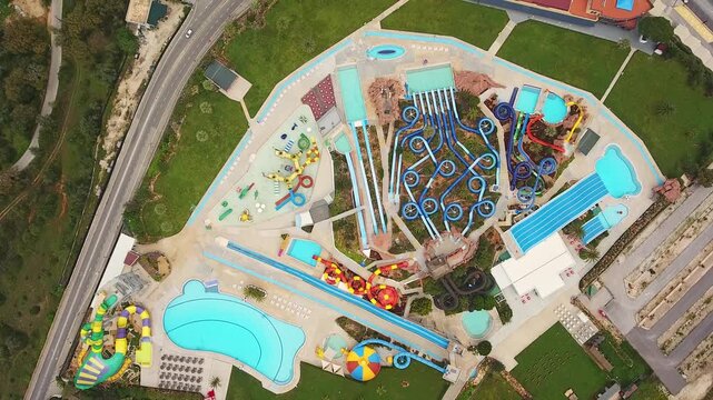 Top down aerial view of a large aquapark with colorful waterslides, swimming pools, and various water attractions. The drone slowly rotates showing the layout of the summer entertainment complex