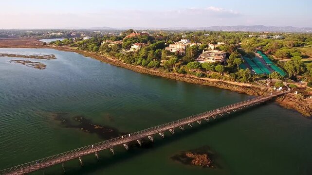Aerial view of the long wooden footbridge connecting quinta do lago to the beach over the ria formosa natural park, with people walking on it during a sunny day in algarve, portugal