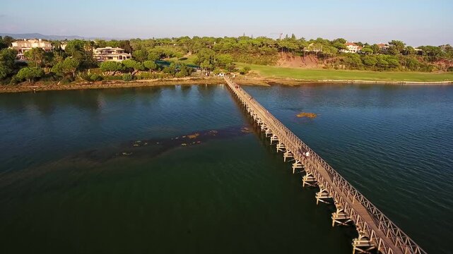 Majestic aerial view showing people walking on the long wooden footbridge at quinta do lago, connecting the mainland to the beach over the ria formosa lagoon in algarve, portugal