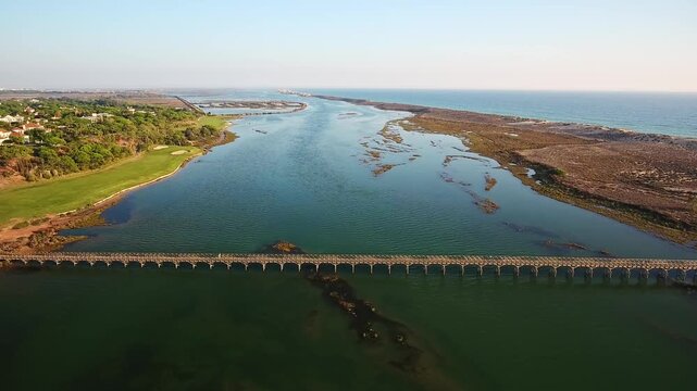 Stunning aerial perspective following a long wooden pedestrian bridge crossing the serene lagoon of ria formosa natural park, connecting a golf course to the ocean beach in algarve, portugal