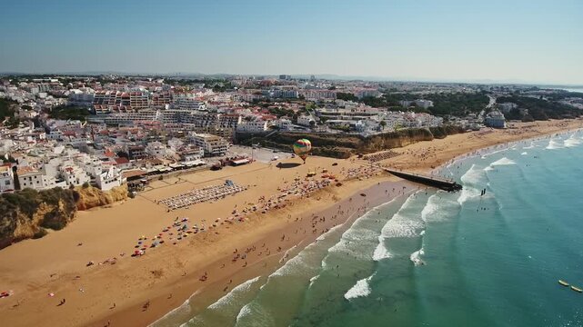 Incredible aerial perspective of the bustling fisherman's beach in albufeira, portugal, featuring tourists enjoying the golden sand, turquoise ocean waves, and a colorful hot air balloon