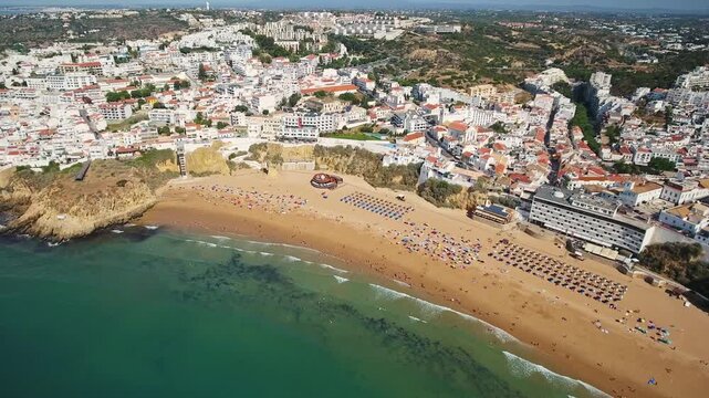 Panoramic aerial view of albufeiras sunlit algarve coast, showing the whitewashed old town, cliffs and bustling praia dos pescadores crowded with summer tourists