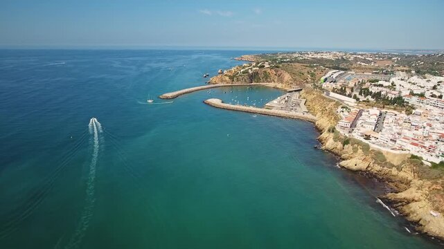 Breathtaking aerial footage of albufeira's stunning coastline, featuring the old town, a marina with a breakwater, and a speed boat cruising on the turquoise atlantic ocean on a sunny day