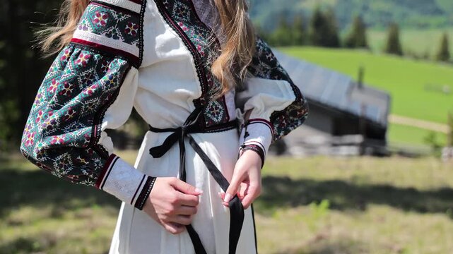 Woman ties a dark sash over an embroidered traditional outfit outdoors, Ethnic style concept highlights handmade ornament history and cultural pride