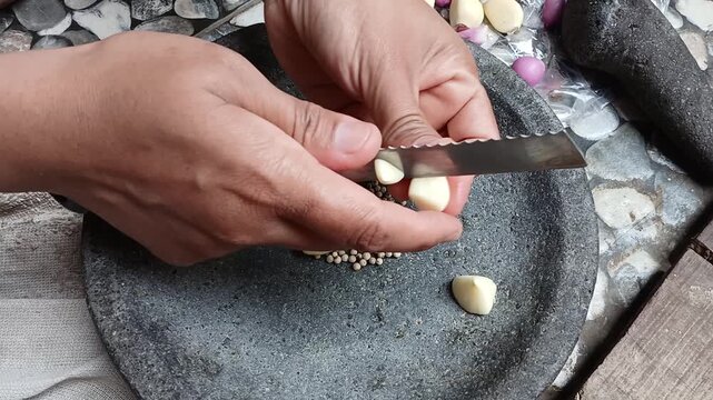 Man slicing fresh garlic cloves and shallots with knife onto traditional stone mortar for Indonesian cooking ingredients preparation