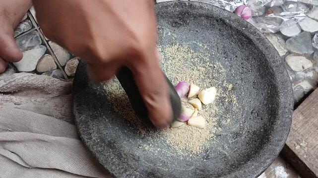 A woman slicing fresh garlic cloves and shallots with knife onto traditional stone mortar for Indonesian cooking ingredients preparation