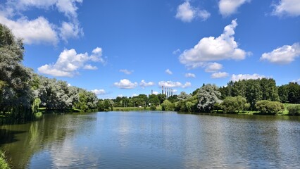 Fototapeta premium Trees grow on the grassy banks of the river. Behind them, factory chimneys rise on the opposite shore. There are ripples and reflections on the water. Sunny summer weather and blue sky with clouds
