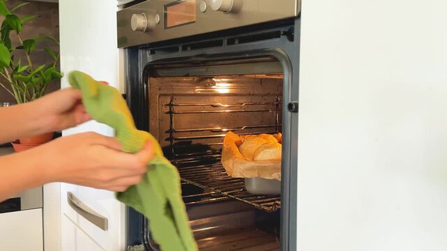 Hands in green oven mitts take tray of freshly baked bread rolls out of oven. Home baking and domestic culinary process concept.