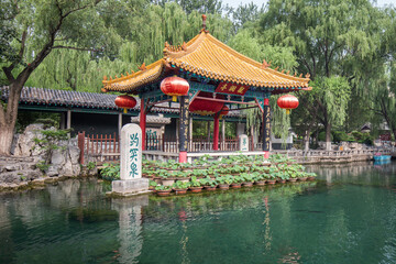 A traditional Chinese pavilion stands by a clear spring pond, with red lanterns and green willows around. © tai