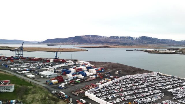 Aerial view over Reykjavik industrial harbor with large vehicle storage lots, warehouses, port facilities and calm coastal water backed by Mount Esja 4K.