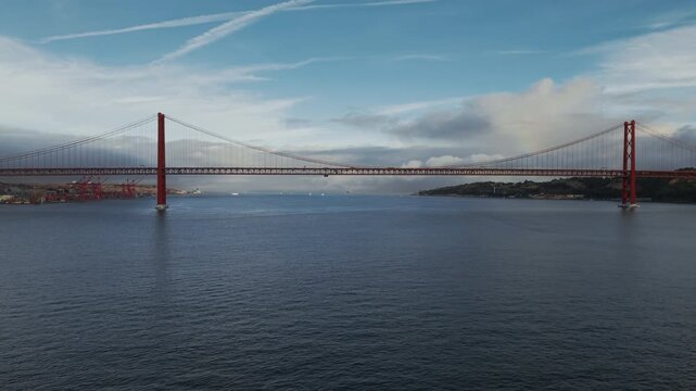 A wide river panorama centers the 25 de Abril Bridge beneath a broken blue and grey cloudscape.
