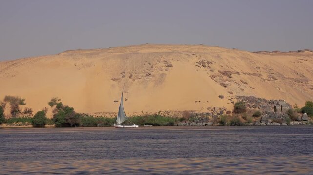 View of a traditional felucca sailing on the Nile River with golden sand dunes in the background in Aswan, Aswan Governorate, Egypt.
