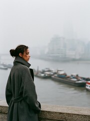 Fototapeta premium Young woman standing on a concrete wall overlooking a body of water. she is wearing a long grey coat and her hair is tied up in a bun.