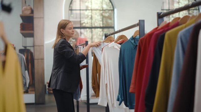 Clothing Store Shopping. Female Retail Assistant Inspects Hanging Apparel, Verifying Quality and Price Tags Before Sorting the New Collection While Shoppers Explore the Contemporary Fashion Boutique.