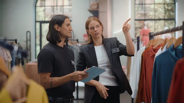 Clothing Store Shopping. Female Shop Manager and a Male Inventory Associate Discuss Stock Data on a Tablet, Sharing Notes and Gesturing at Displays While Coordinating the Next Restocking Round.