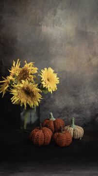 Autumn still life with a bouquet of yellow sunflowers in a glass vase and cozy crocheted pumpkins on a dark background.