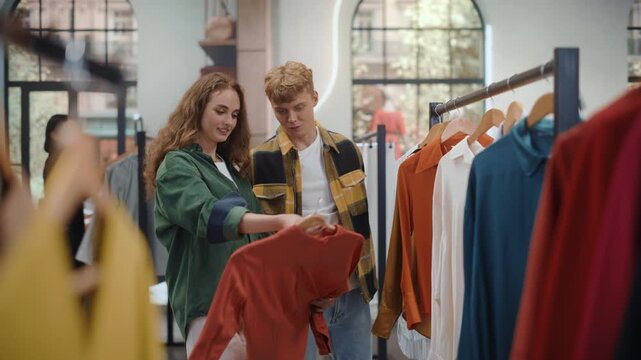 Clothing Store Young People Shopping. Happy Couple Browsing Racks Together, Discussing Choices and Holding up a Colorful Garment While Smiling, Enjoying a Relaxed Weekend Retail Shopping Date.