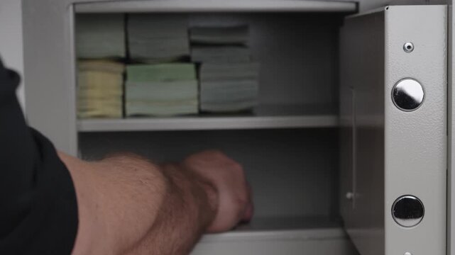 Male hand placing black pistol inside open metal safe box filled with stacks of paper currency. Close up view on security storage, wealth protection and hidden firearm.
