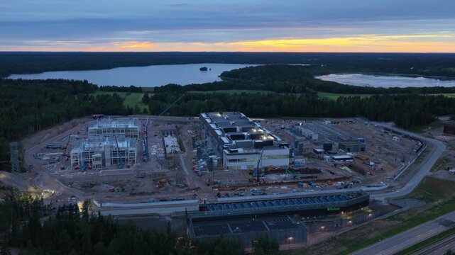 Aerial view of data center construction site with cranes and industrial buildings surrounded by forest and lake at sunset in Espoo, Uusimaa, Finland.
